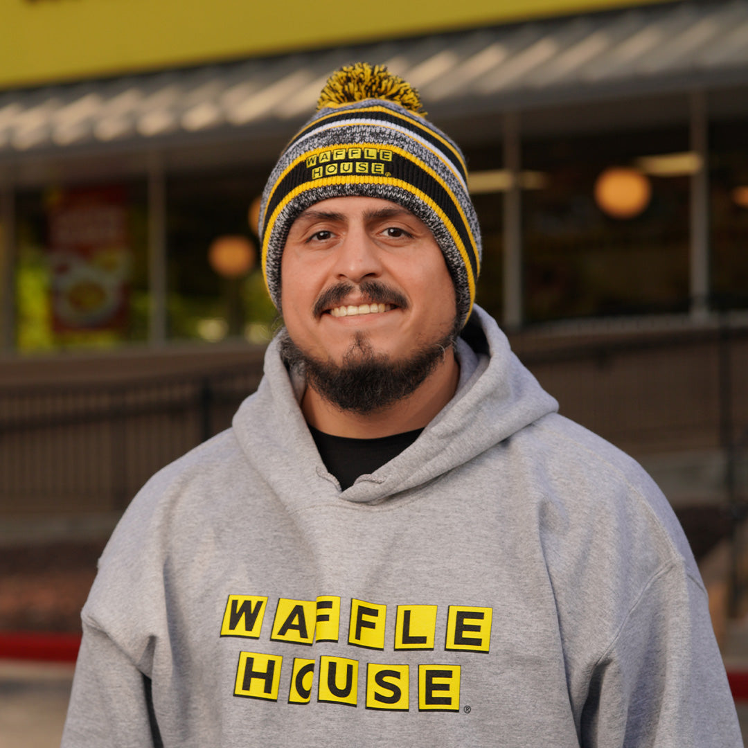 black, yellow and grey striped beanie with pom on top and Waffle House logo on cuff being worn by a man standing in front of a Waffle House