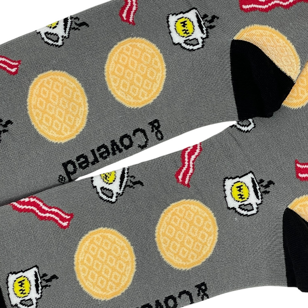 Close up view of Pair of gray socks with black trim and images of waffles, bacon and coffee and the Waffle House logo.