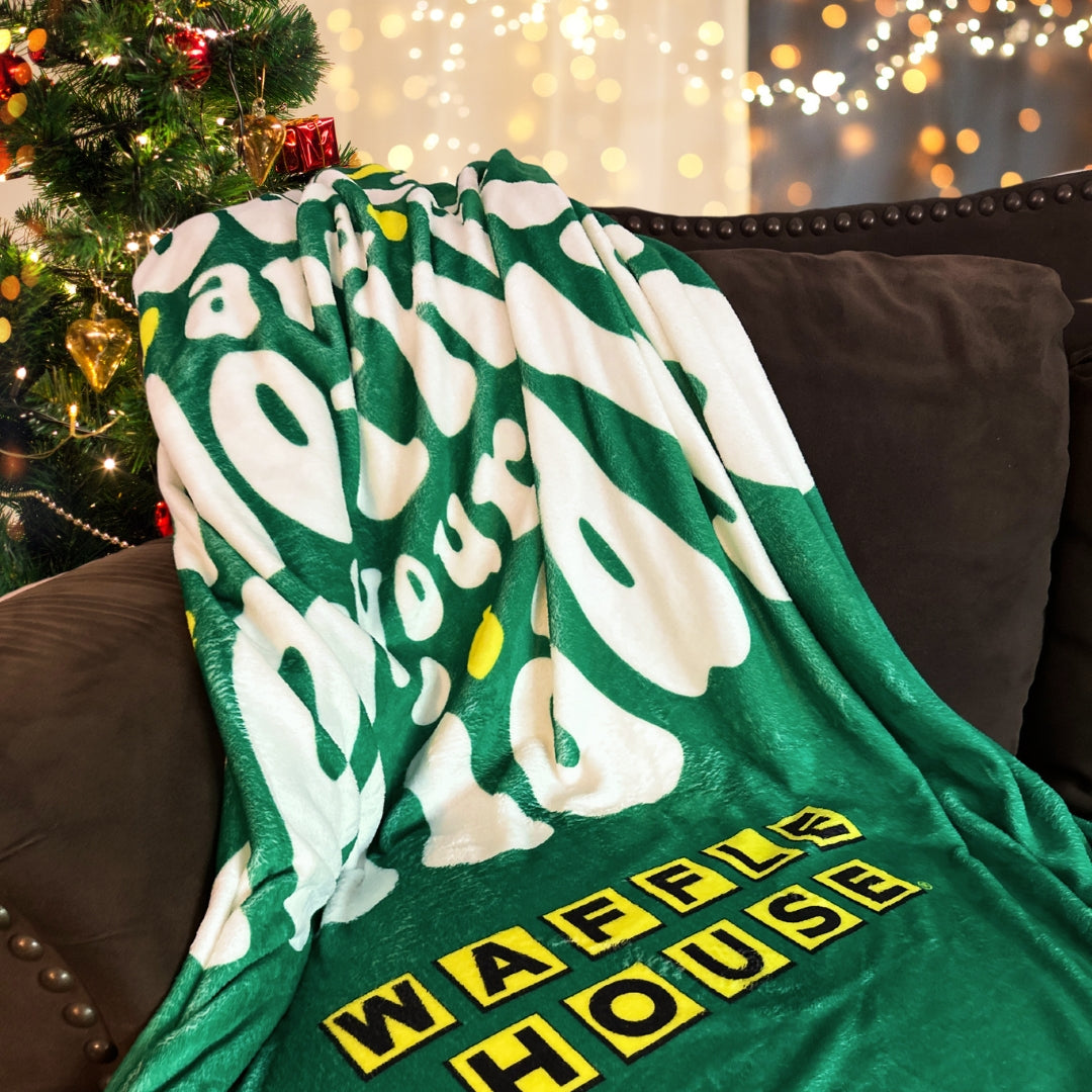 green Waffle House holiday blanket draped over a brown couch with Christmas tree in the background