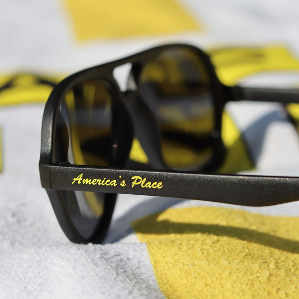 Black sunglasses with America's Place written in yellow on the arm, laid out on a towel