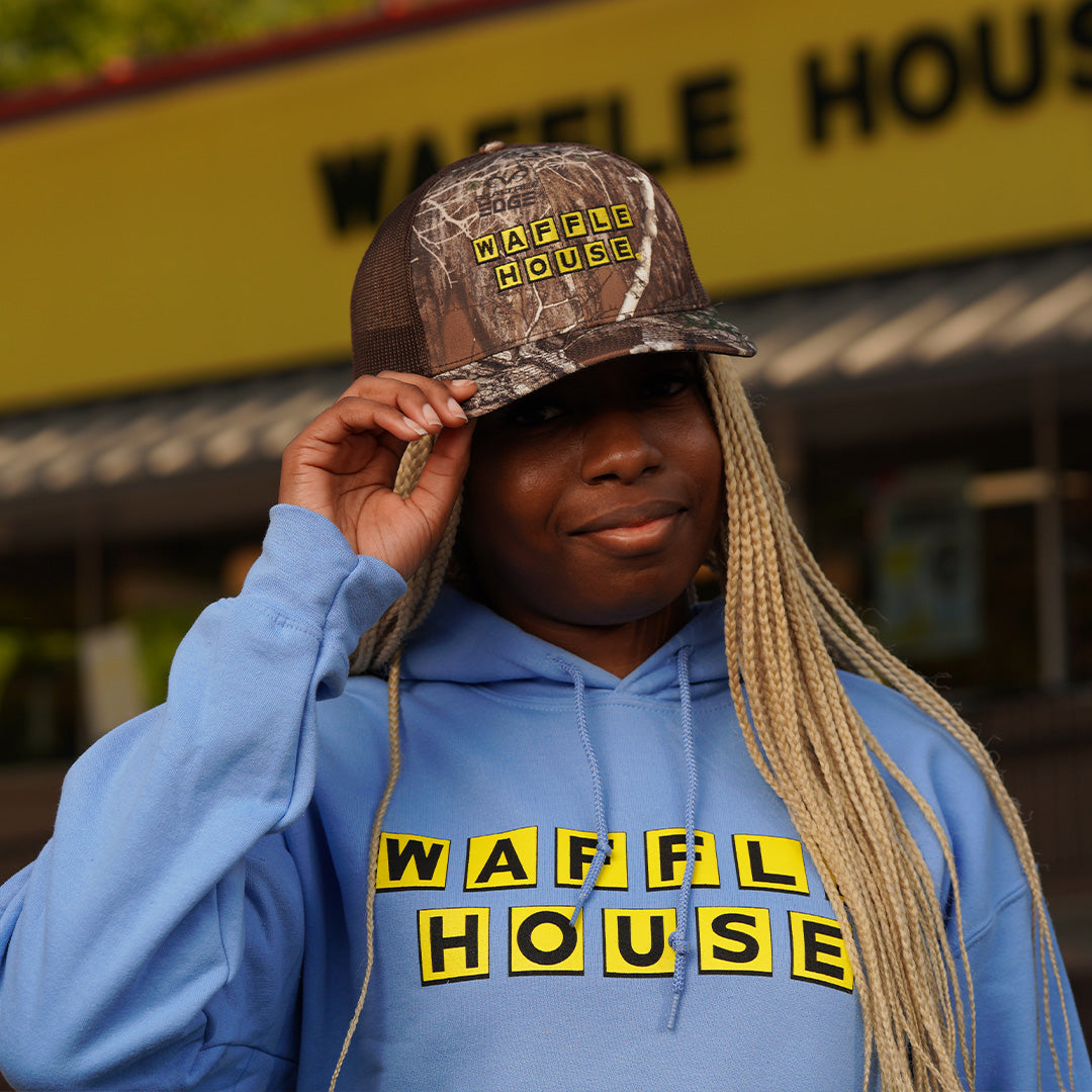 Woman wearing camo trucker hat with a brown mesh back and the Yellow and black Waffle House logo on the front standing in front 