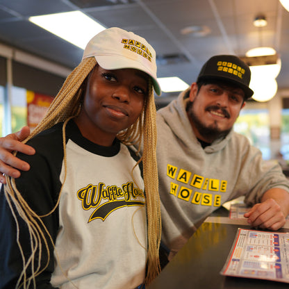 Man and woman inside Waffle House sitting with Waffle House hats on. Woman wearing Waffle House baseball tshirt. Man wearing Waffle House hoodie.