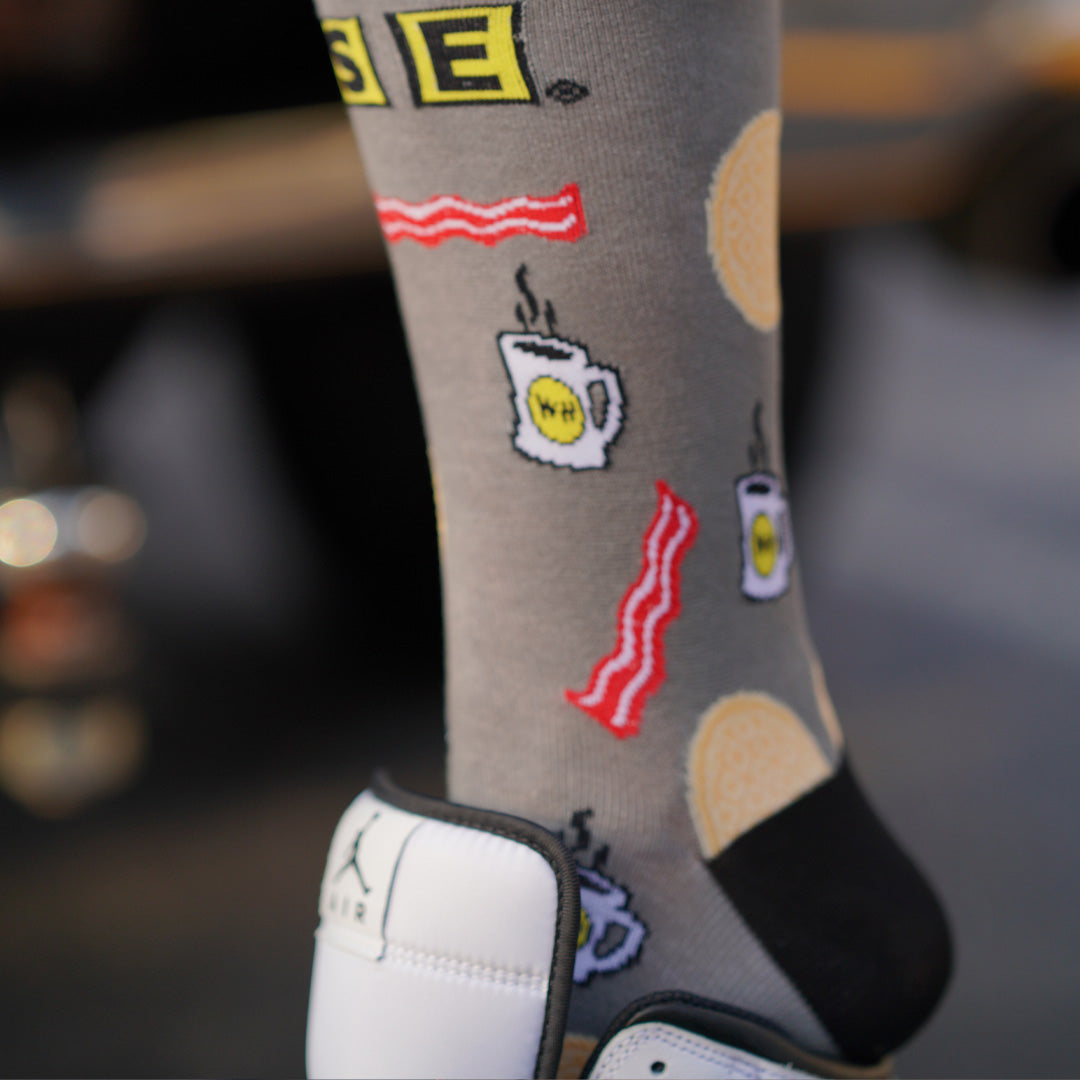 Person wearing Waffle House dress socks onto their leg. Showing off Waffle House logo, waffles, bacon, coffee displayed on the socks.