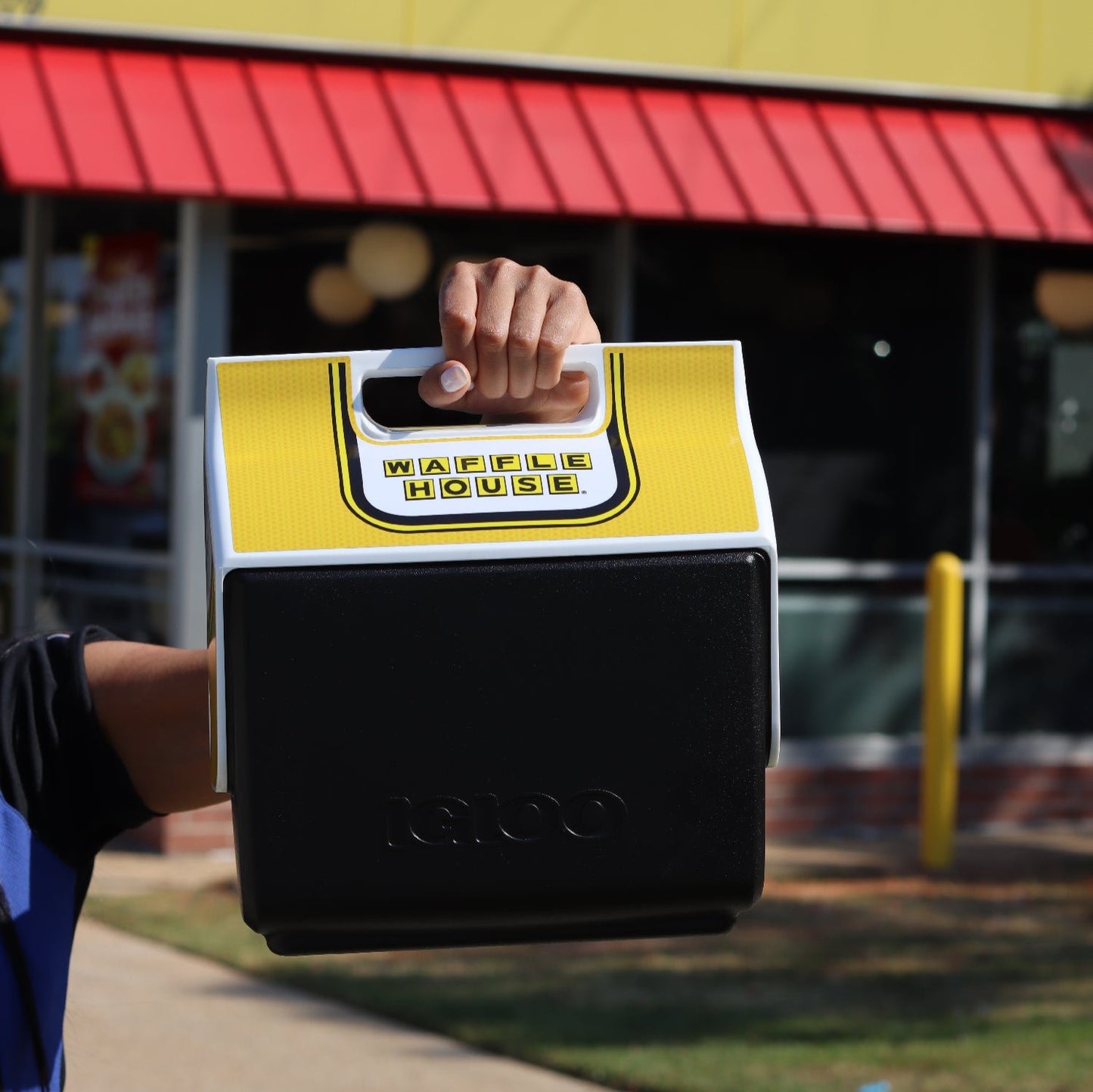 Black and Yellow Igloo cooler with the Waffle House logo held up in front of a Waffle House store