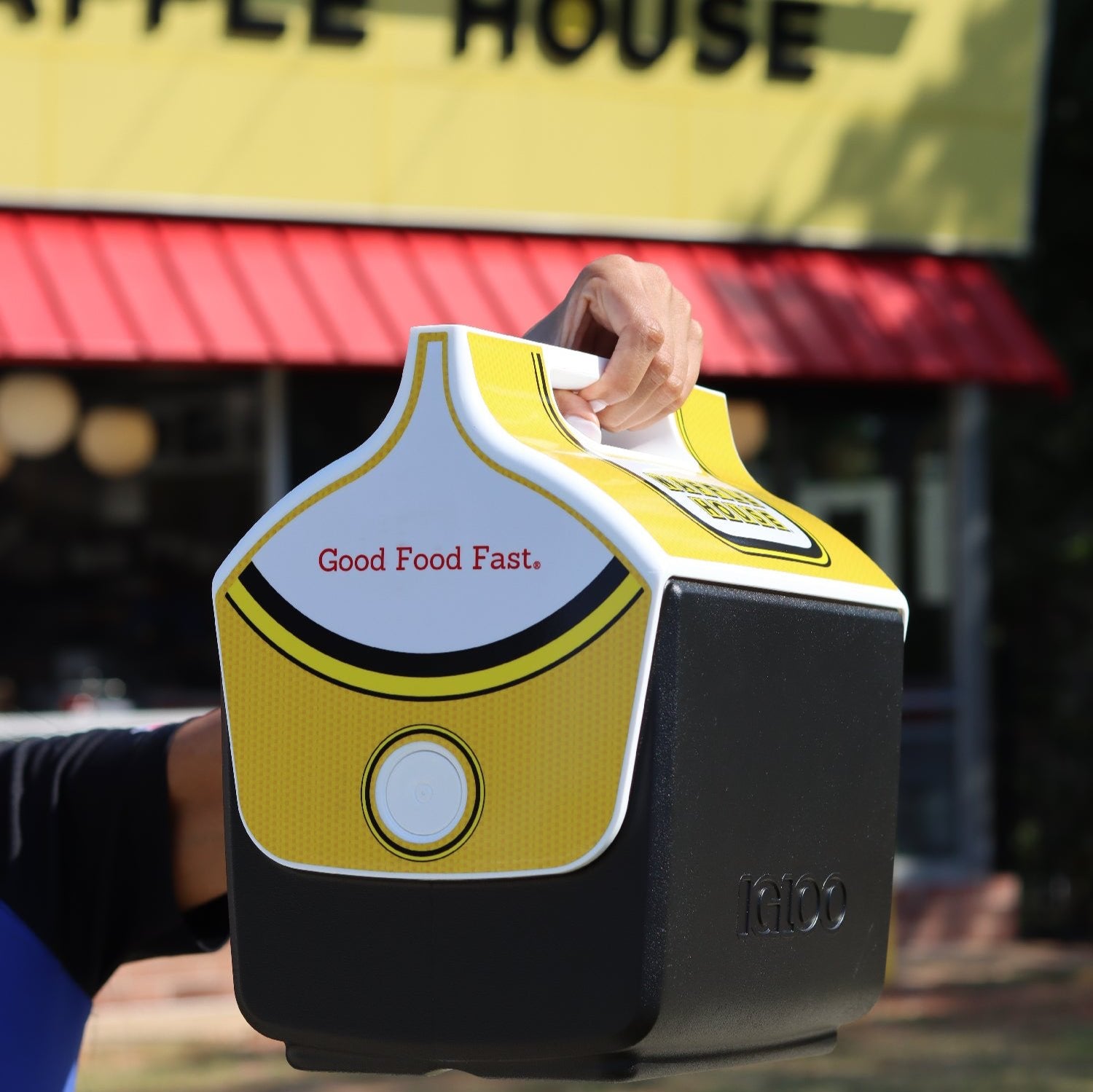 Black and Yellow Igloo cooler with the Waffle House logo  and Good Food Fast printed on the side held up in front of a Waffle House store